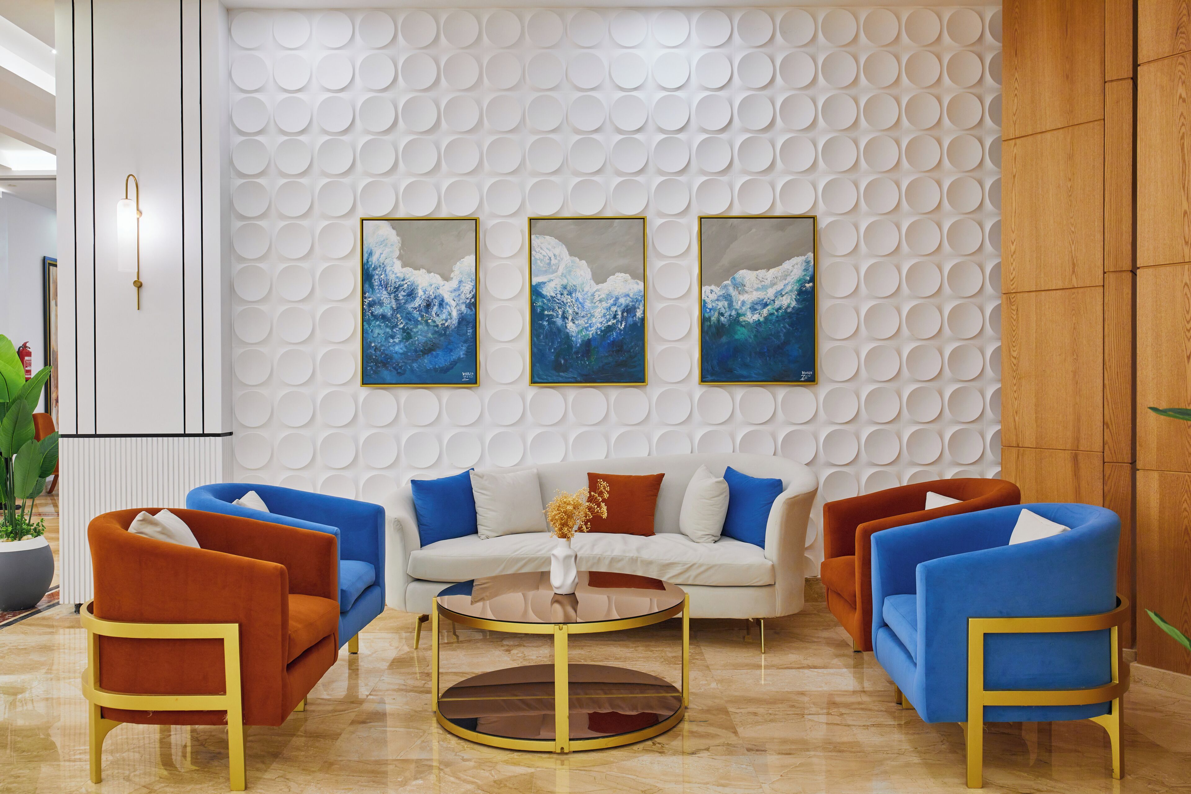 Una sala moderna con un sofá blanco, sillones azul y naranja, una mesa de centro de vidrio y tres cuadros en una pared con textura.