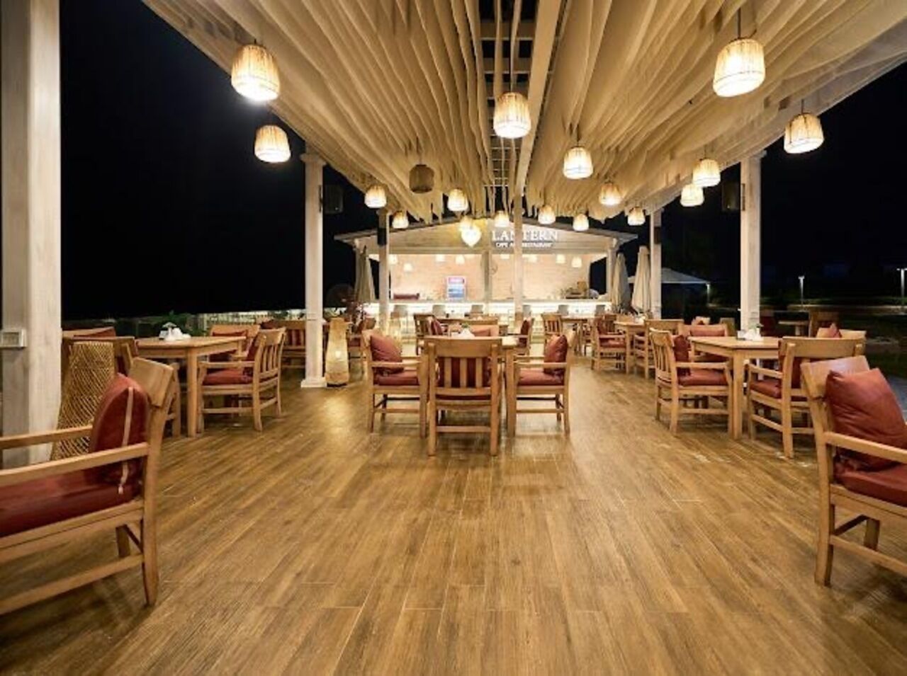 Un espace de restauration en plein air avec des tables et des chaises en bois, une partie couverte agrémentée de guirlandes lumineuses, et un restaurant en arrière-plan.