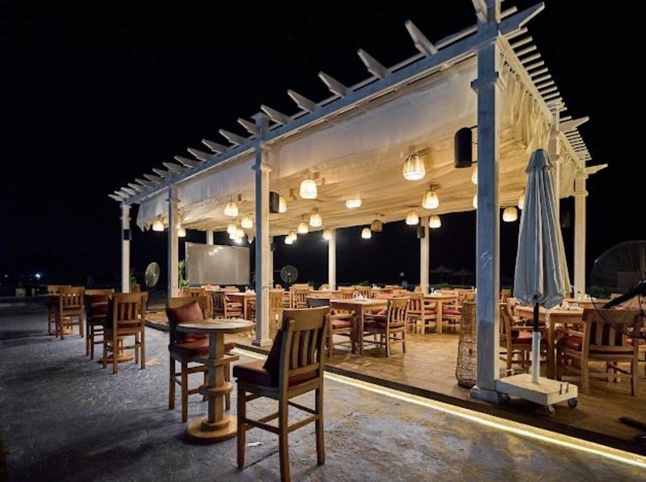 Un restaurante al aire libre con mesas y sillas de madera, cubierto por una pérgola con luces colgantes.