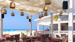 Een terras met tafels en stoelen, uitzicht op het strand, en een gebouw met een bord waarop 'LANT CAFE AND RESTAURANT' staat.