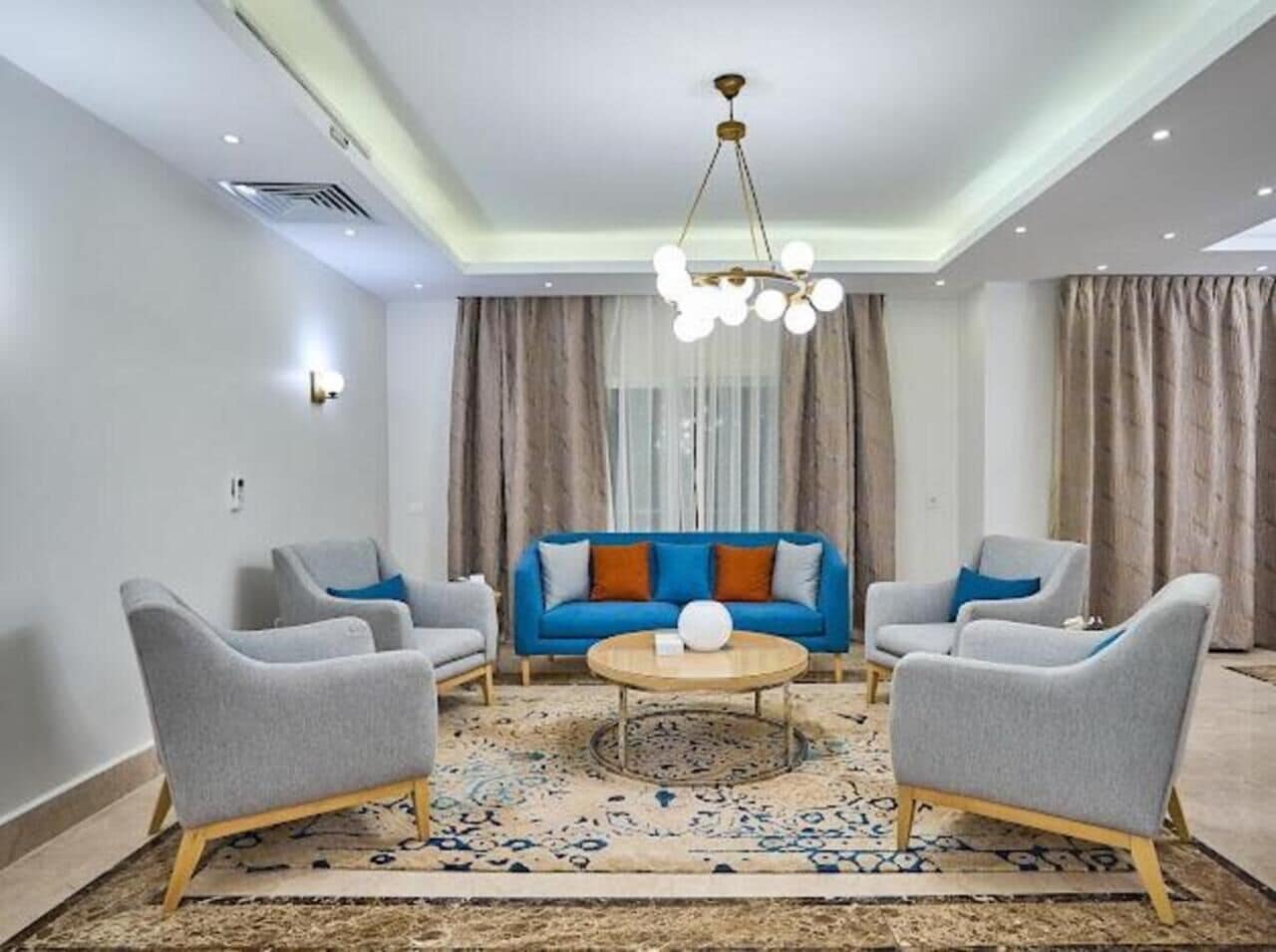 Una sala de estar con un sofá azul, sillones grises, una mesa de centro redonda y una araña.