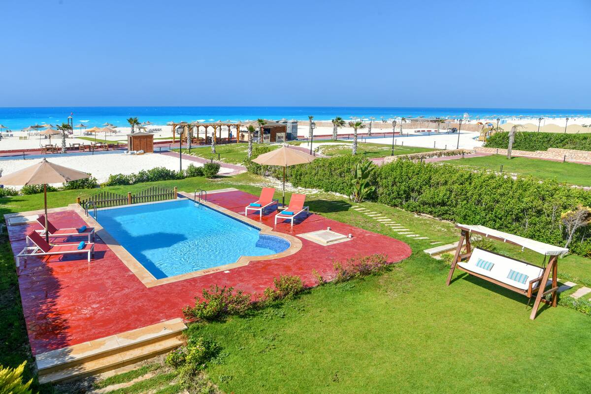 Un espace piscine doté d’un sol rouge, entouré de pelouse et offrant une vue sur la plage.
