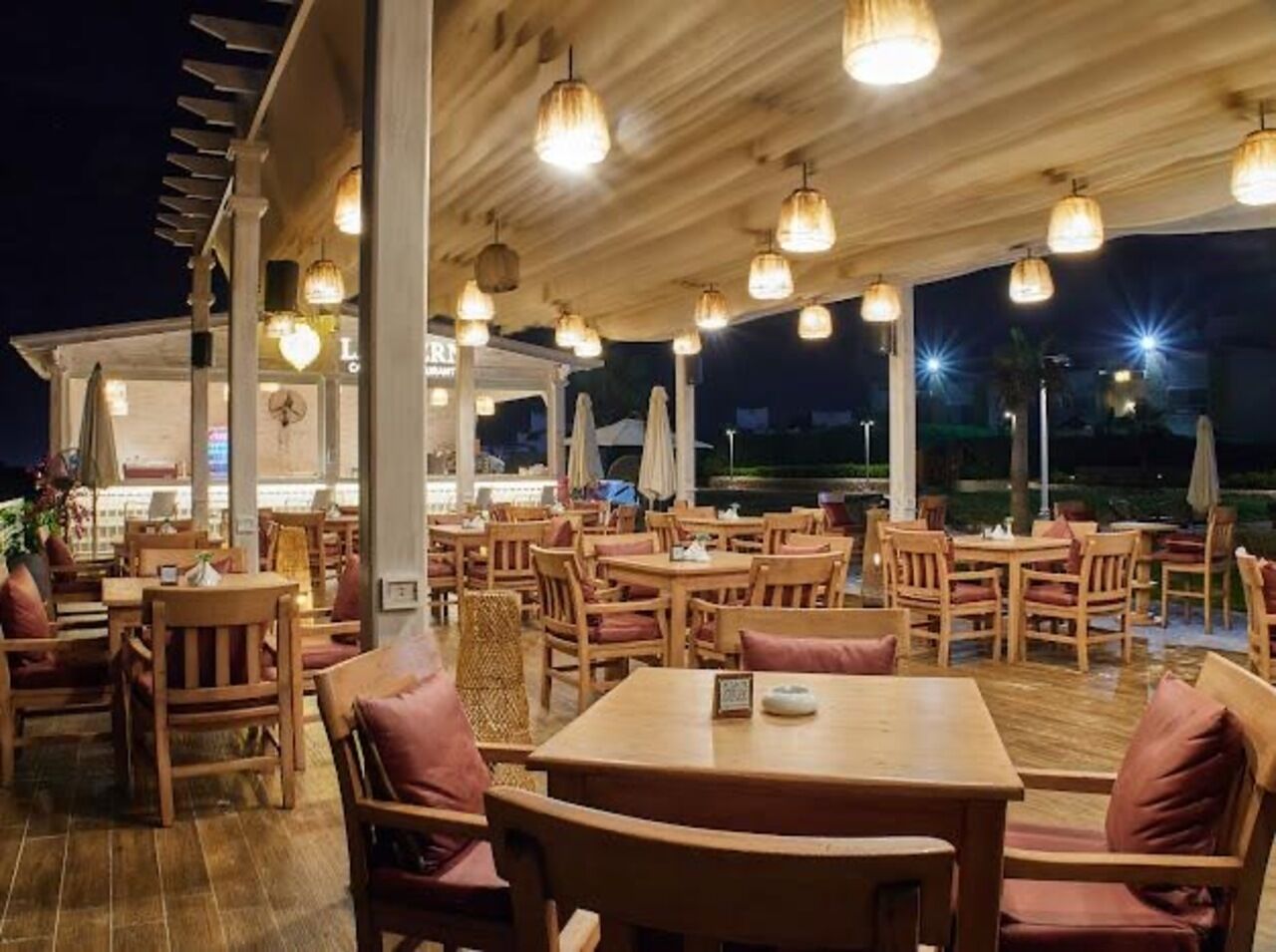 Un restaurante al aire libre con mesas y sillas de madera, iluminación colgante y vista al cielo nocturno.
