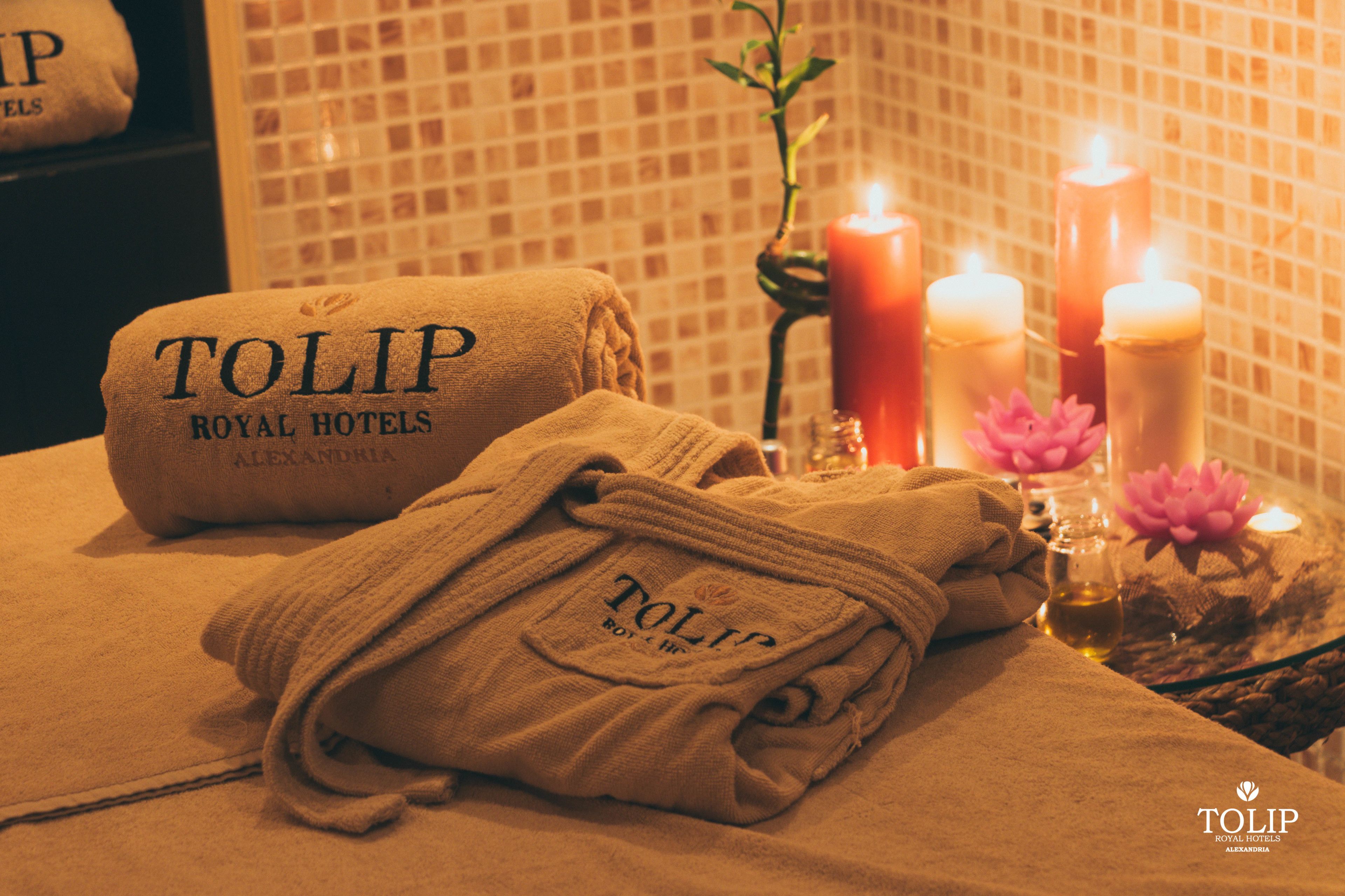 Siististi taitettu pyyhe, jossa on teksti ”TOLIP ROYAL HOTELS ALEXANDRIA”, sytytetty kynttilä ja lootuksenkukka pöydällä.