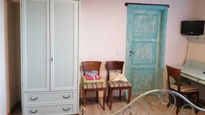 Une pièce avec une grande armoire blanche, une porte de couleur turquoise, deux chaises en bois, un cadre de lit en métal et un téléviseur fixé au mur.