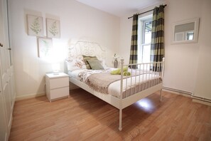 A bedroom with a white metal bed frame, a bedside table, a nightstand, and a window with curtains.