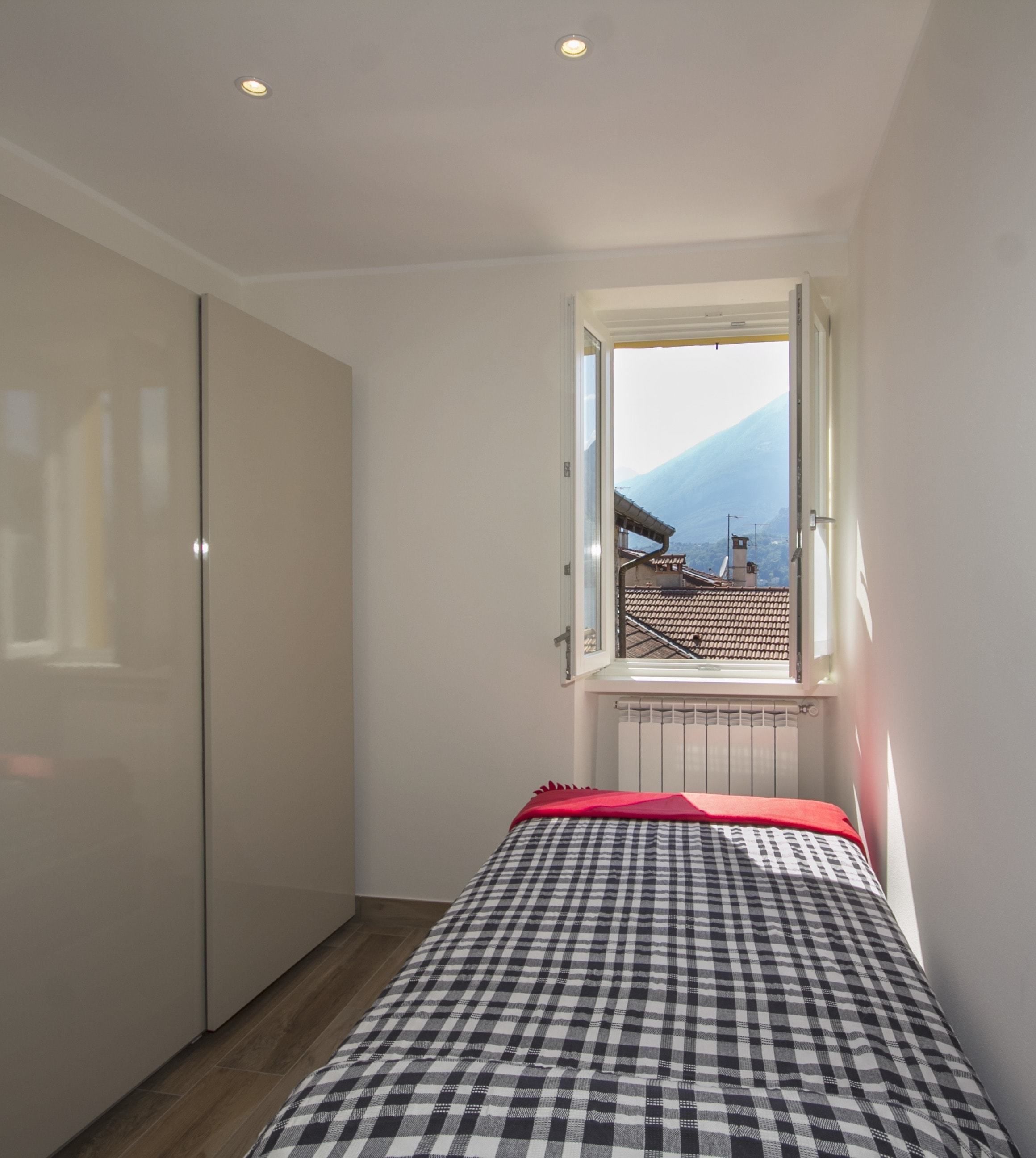 Un dormitorio con edredón a cuadros, una manta roja y una ventana con vistas a una montaña y un pueblo.