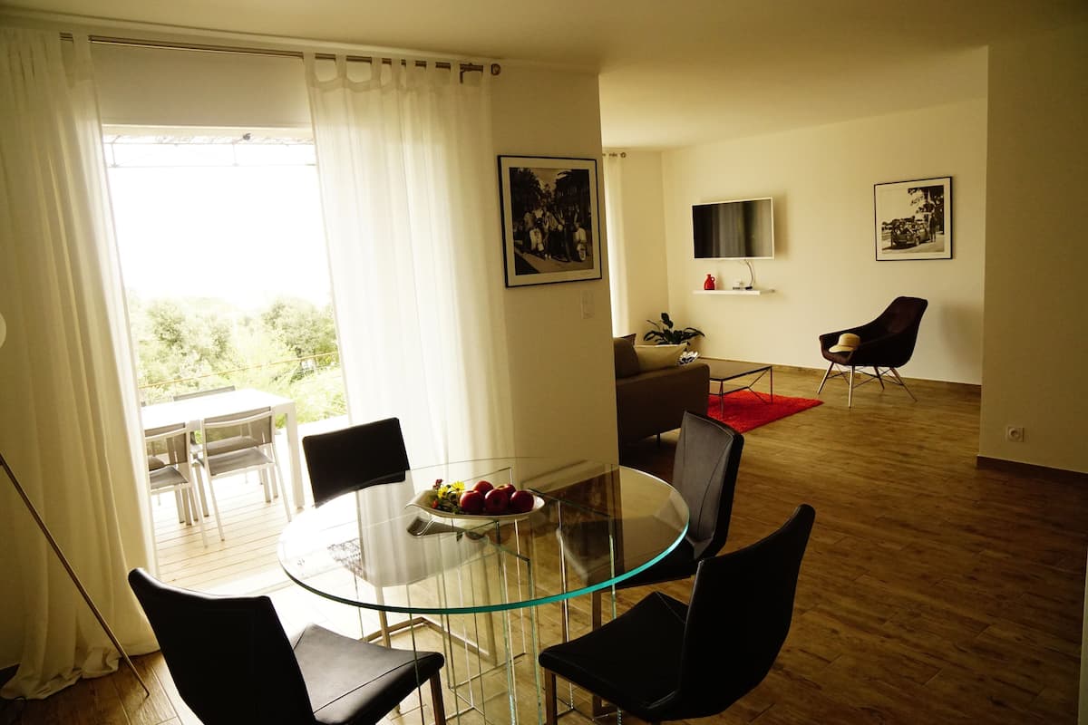 A modern living room with a glass dining table, black chairs, a red rug, and a TV on the wall.