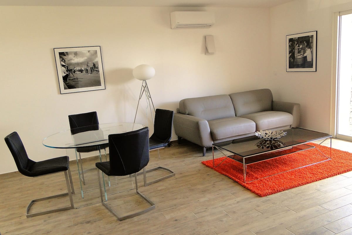 A modern living room with a glass dining table, black chairs, a grey sofa, and a coffee table with a red rug.
