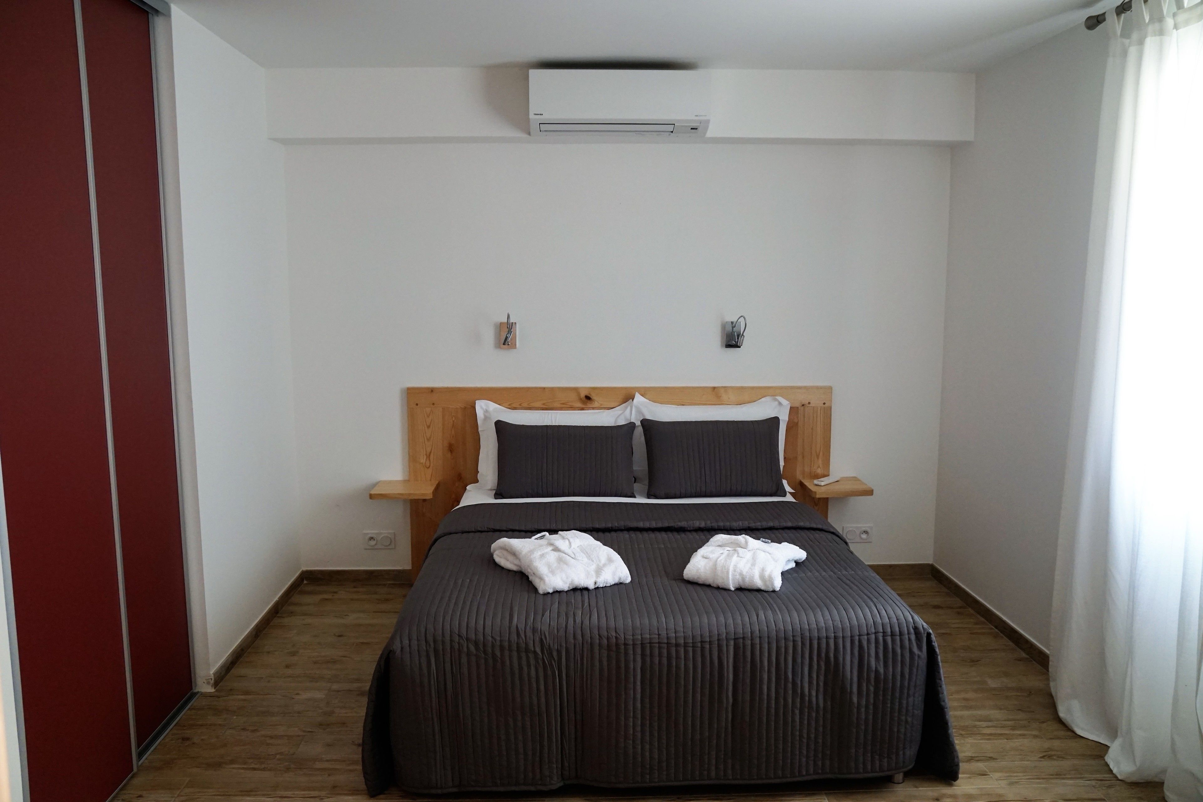 Une chambre d’hôtel avec un lit, une tête de lit en bois, un climatiseur fixé au mur et une porte rouge.