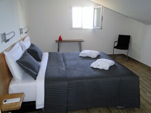 Una cama bien hecha con un edredón oscuro, almohadas blancas y dos albornoces blancos. Hay una mesita de madera con una botella roja encima, una silla negra y una ventana con persianas.