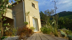 A wooden deck leading to a beige building with a door and window, surrounded by greenery and a hilly landscape. - Suites in Erbalunga (Brando)