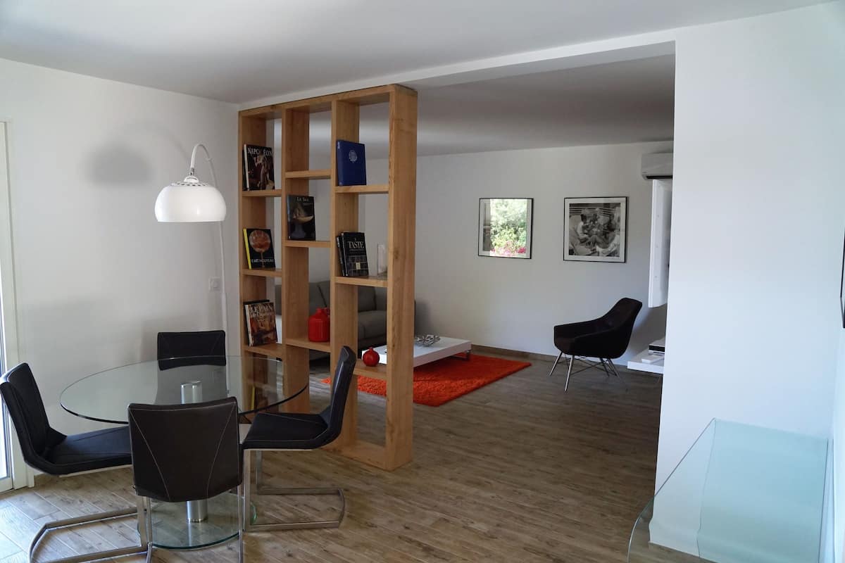 A modern living room with a glass dining table, black chairs, a wooden bookshelf, and a red rug.