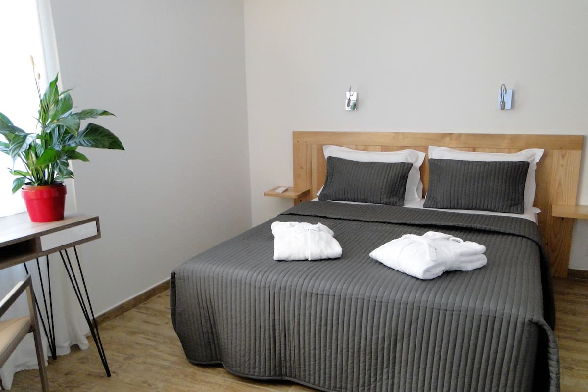 A neatly made bed with gray bedding, two pillows, and two towels. There is a wooden headboard, a bedside table with a red plant, and a white chair.