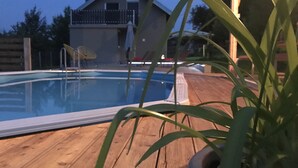 Une piscine avec une terrasse en bois, une maison en arrière-plan et une plante au premier plan.