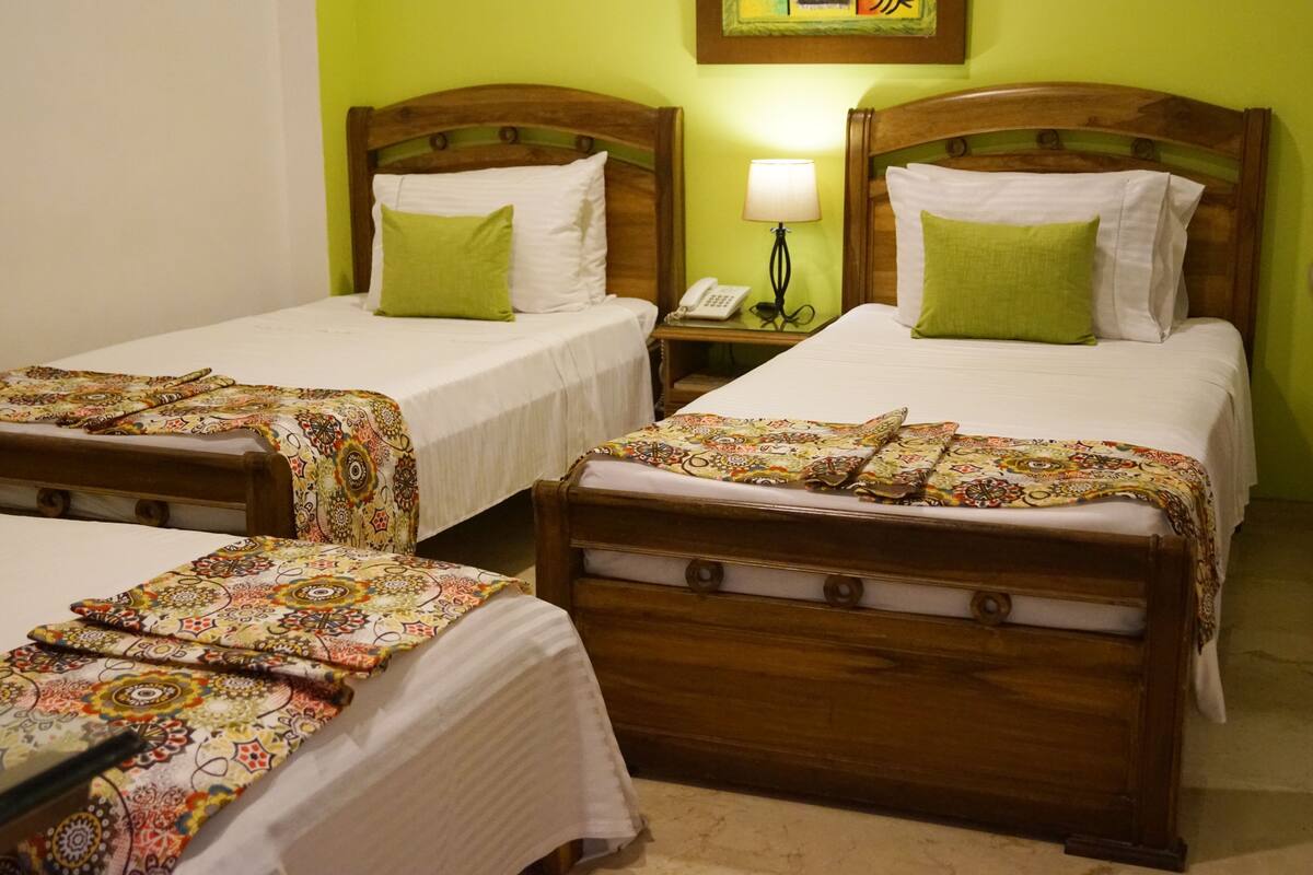 A hotel room with two wooden beds, each with a green pillow and a patterned bedspread. There is a nightstand with a lamp and a phone between the beds.
