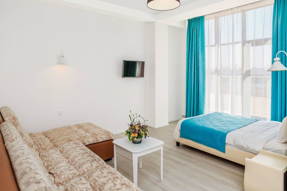 A modern hotel room with a bed, a sofa, a small table with a vase of flowers, a TV, and large windows with curtains.