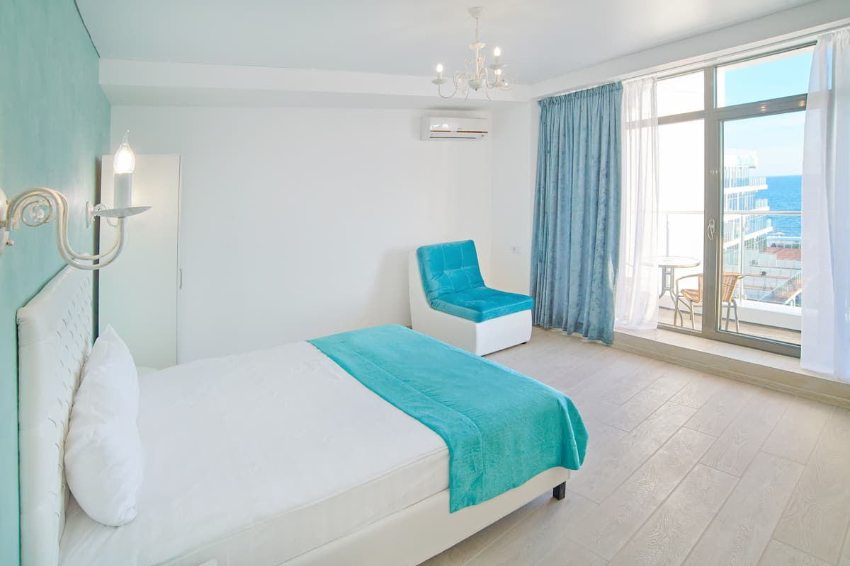 A hotel room with a bed, a chair, a balcony with a view of the sea, and a chandelier.