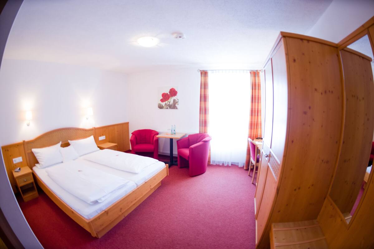 A hotel room with a large bed, a wooden wardrobe, a small table, two red chairs, and a window with curtains.