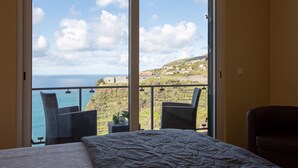 Habitación de hotel con un ventanal grande que ofrece vista al mar y a una colina.