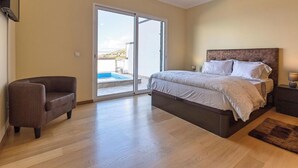 Un dormitorio con cama, una silla, una mesita de noche, una lámpara y vista a una piscina a través de una puerta corredera.