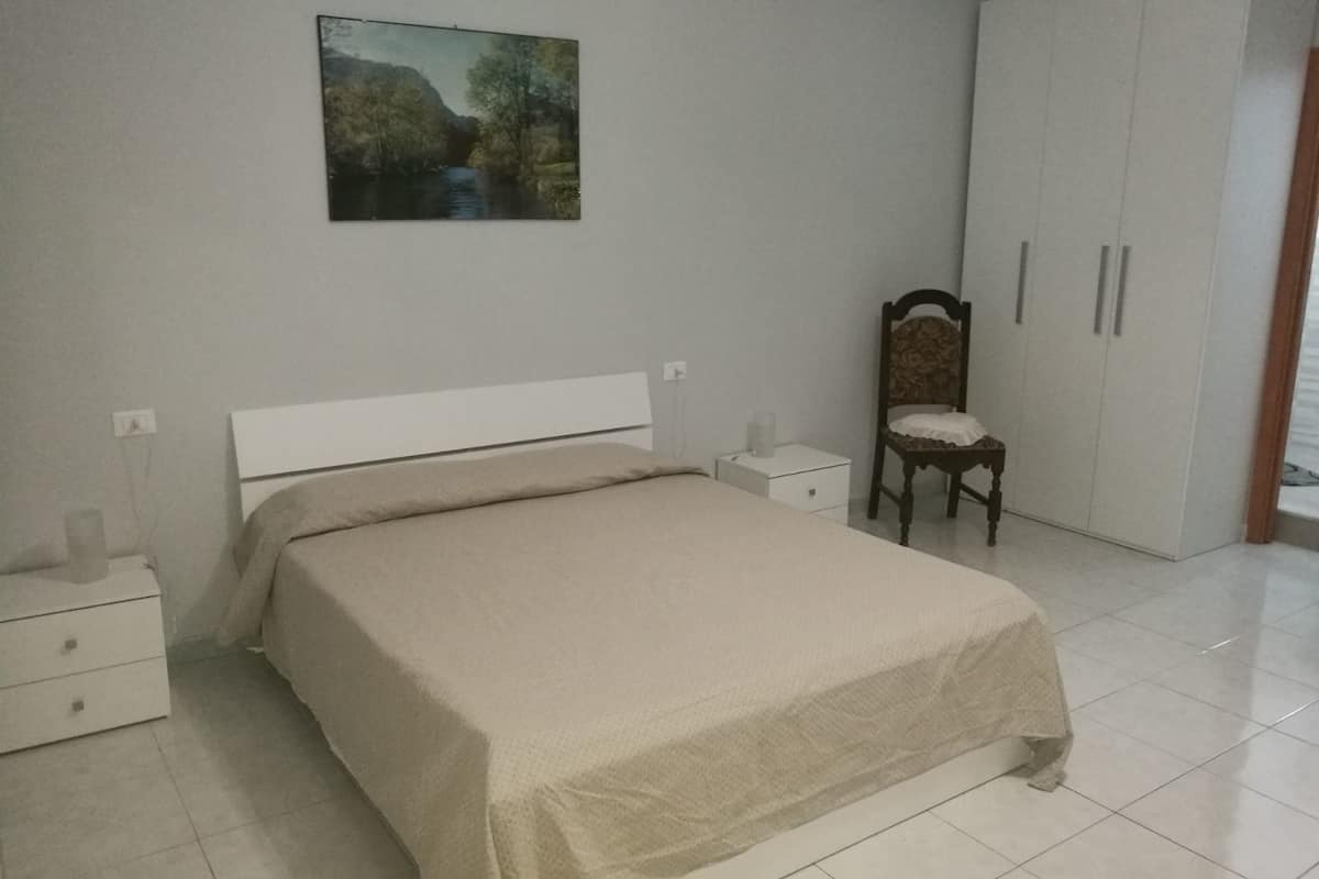 Een slaapkamer met een bed, nachtkastje, stoel, kledingkast en een schilderij aan de muur.