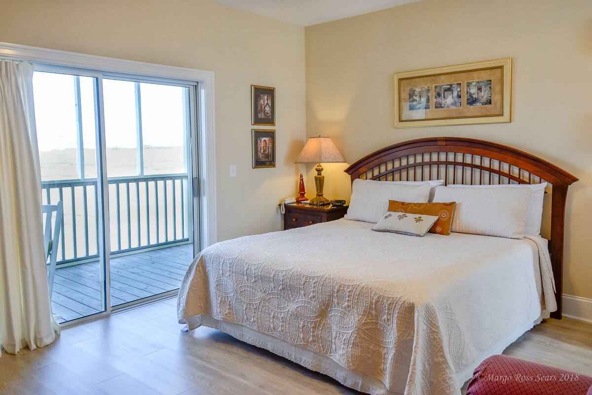 A bedroom with a large bed, a nightstand, a lamp, and a view through a sliding glass door to an outdoor balcony.