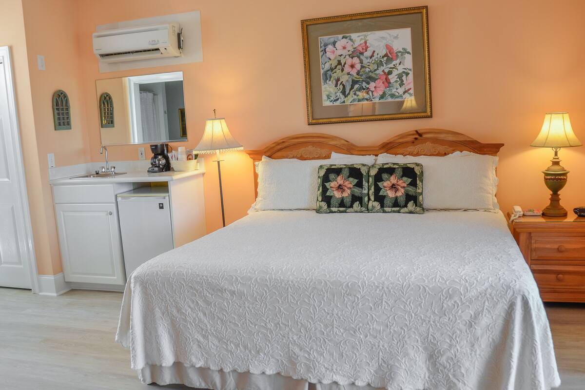 A bedroom with a bed, bedside lamps, a nightstand, a dresser, a desk with a sink, and a mirror.
