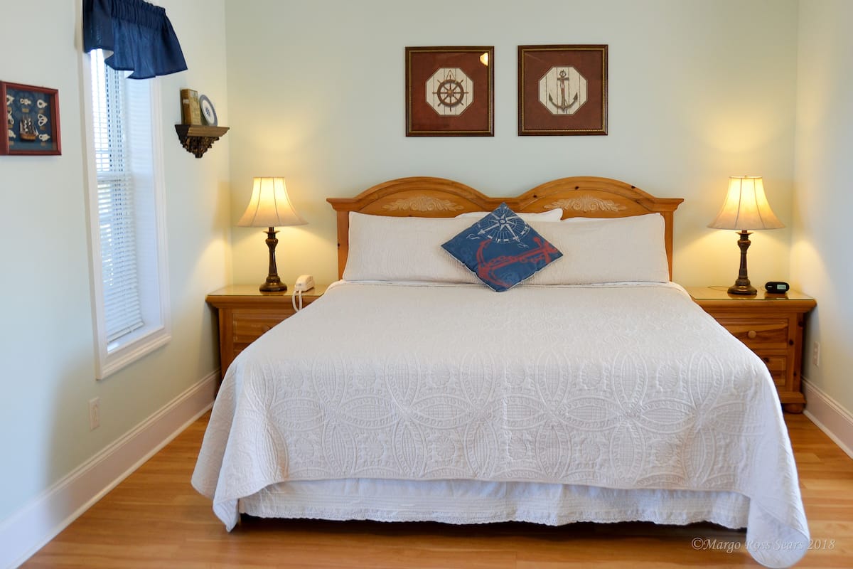 A large bed with a white quilt, two bedside tables with lamps, a window with blinds, and two framed nautical-themed pictures on the wall.