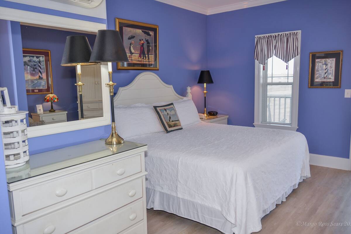A bedroom with a white bed, two nightstands, a dresser, a large mirror, and a window with curtains.