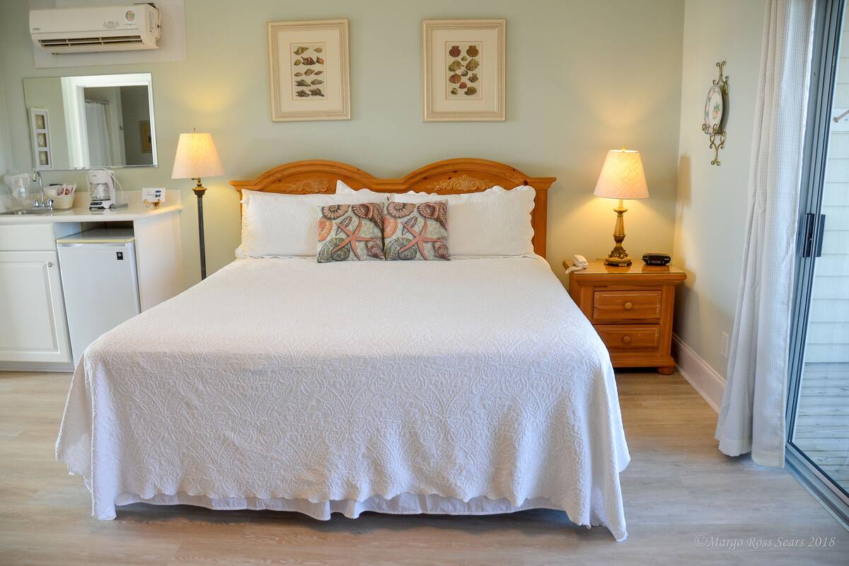 A neatly made bed with a white bedspread, two decorative pillows, a bedside table with a lamp, and a small nightstand.