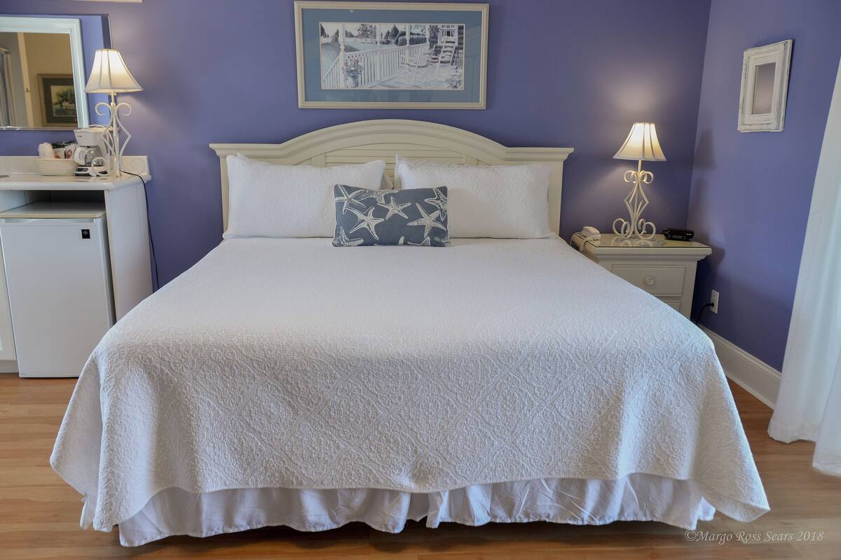 A neatly made bed with a white headboard, a bedside table with a lamp, and a small desk with a lamp and a telephone.
