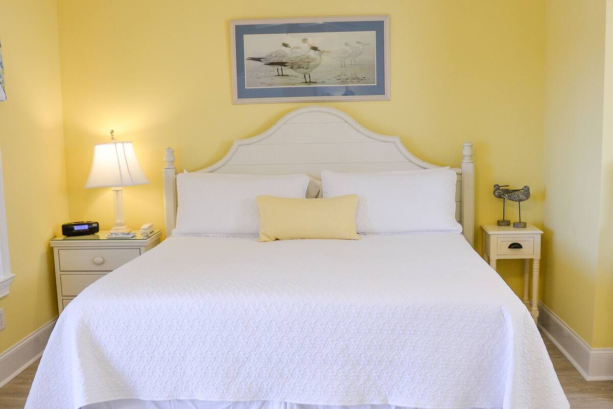 A neatly made bed with white linens and a ruffled skirt, a bedside lamp, a nightstand with a radio, and a framed picture of birds on the wall.