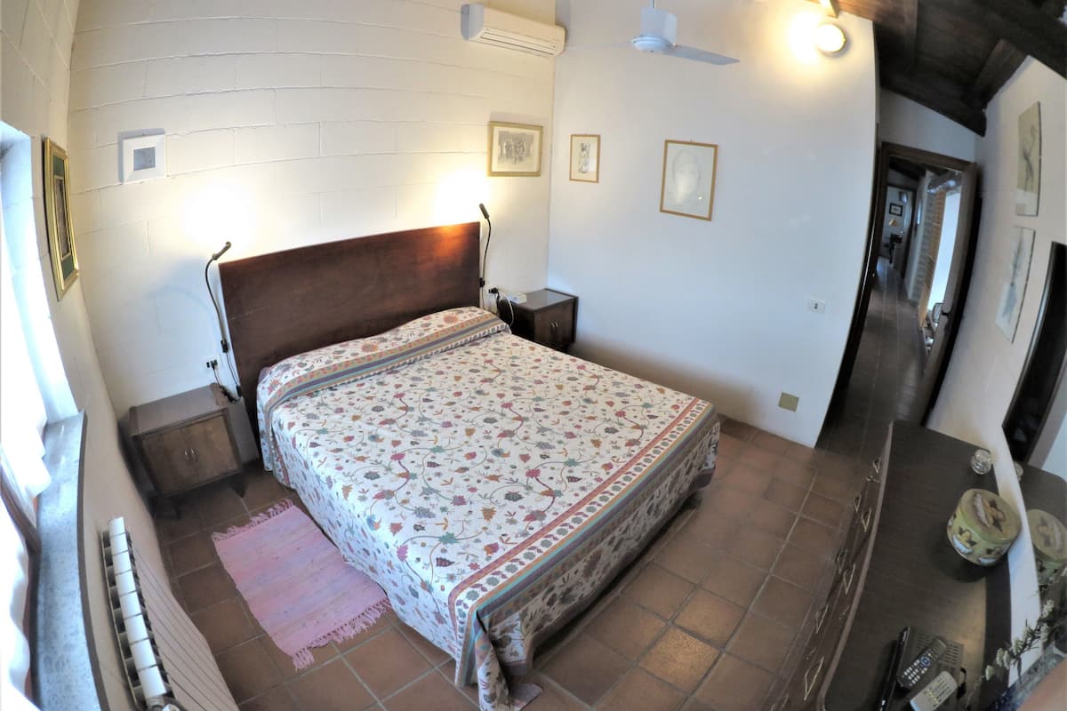 Une chambre à coucher avec une tête de lit en bois, une couvre-lit à motifs, une table de chevet et une commode, ainsi qu’un climatiseur fixé au mur.