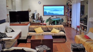 Una sala de estar acogedora con un sofá, televisor, mesa con sillas, estantería y ventana con vista al exterior.