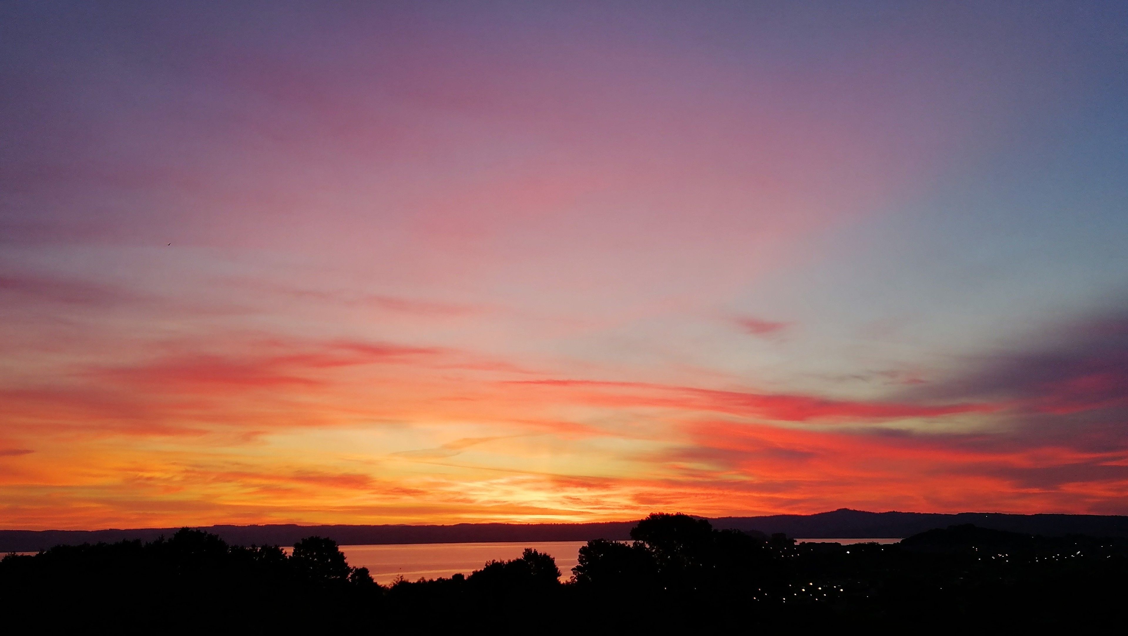 A sunset over a body of water with silhouettes of trees and distant hills.