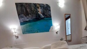 A hotel room with a bed, a wall-mounted TV displaying a coastal scene, and a mirror. - Affittacamere Maristella (Cagliari)
