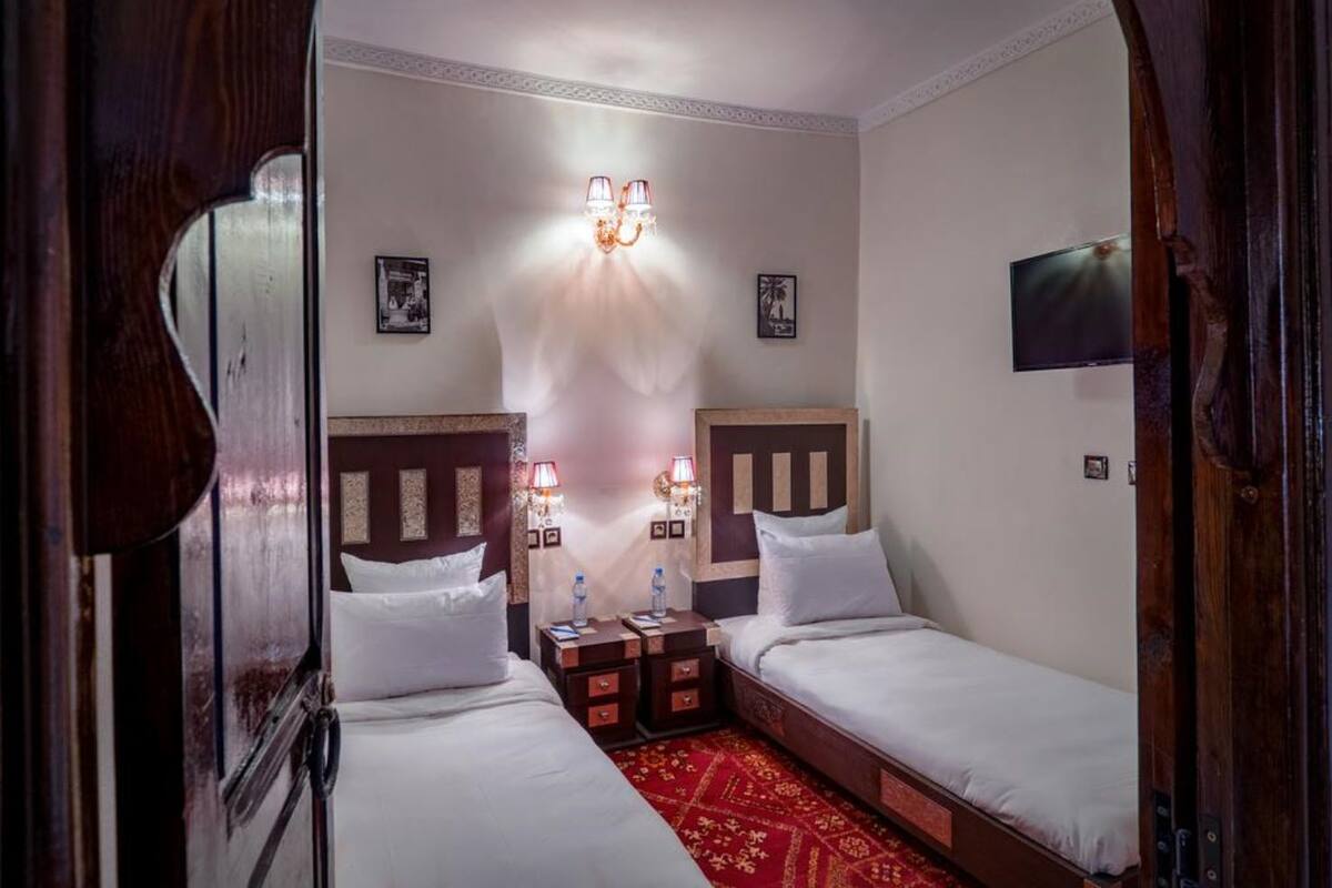 A hotel room with two single beds, a red patterned carpet, a wall-mounted TV, and a chandelier.