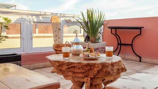 Una mesa de madera con una tetera, vasos y aperitivos en una terraza con vistas a edificios y una palmera.