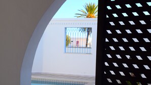 A view through a window with a pool and a white building with a blue door and a window with a metal gate. - House Dar Mamina (Zarzis)