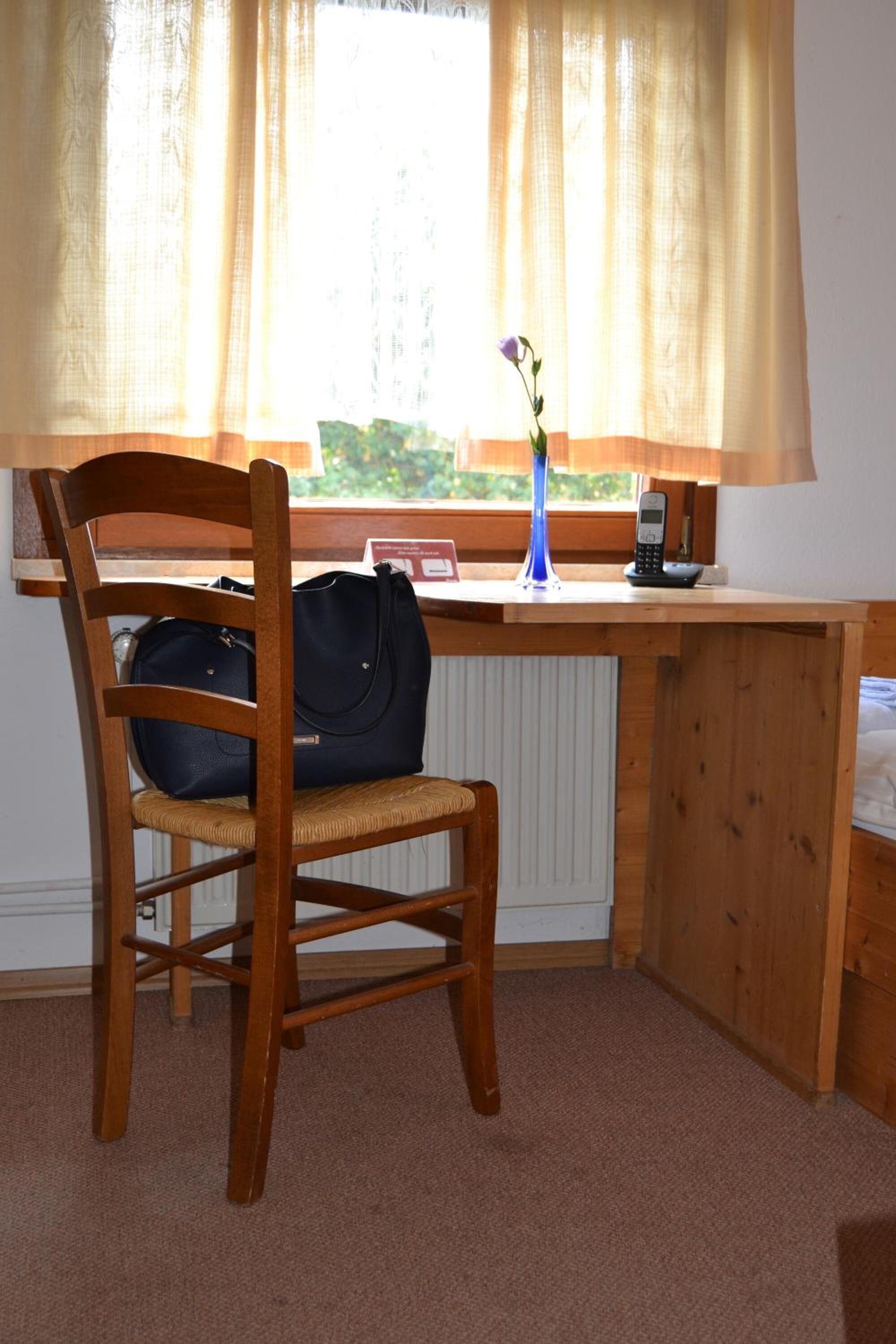 Un bureau en bois avec une chaise, un vase bleu avec une fleur, une lampe et un téléphone portable.