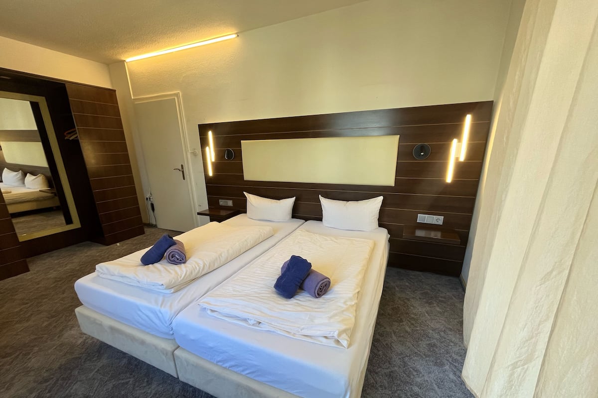 Uma cama de casal com lençóis brancos e duas toalhas dobradas, uma cabeceira de madeira, uma mesa de cabeceira e um espelho.