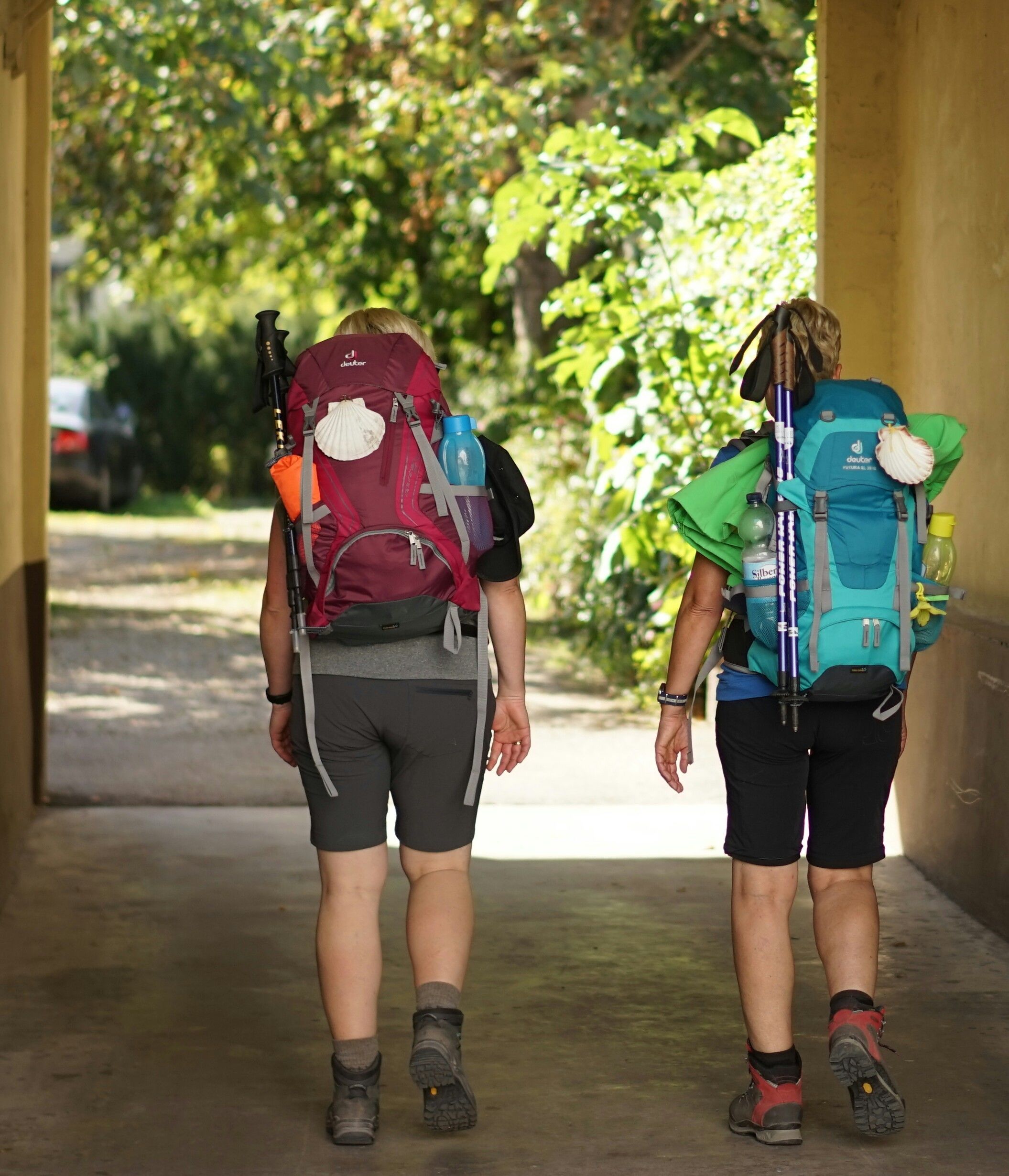Dos personas caminando por un pasillo cubierto con mochilas, una roja y otra azul, sobre un piso de concreto.