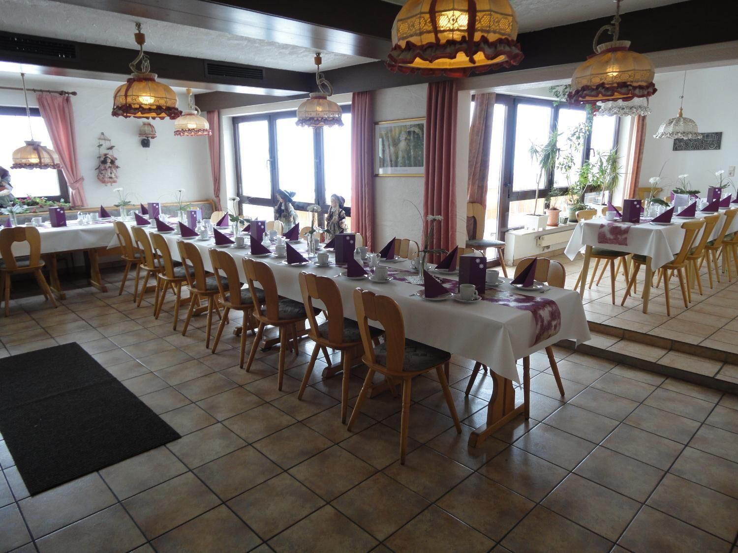Un espace repas avec plusieurs tables dressées pour le service, des chaises en bois et des lampes suspendues.
