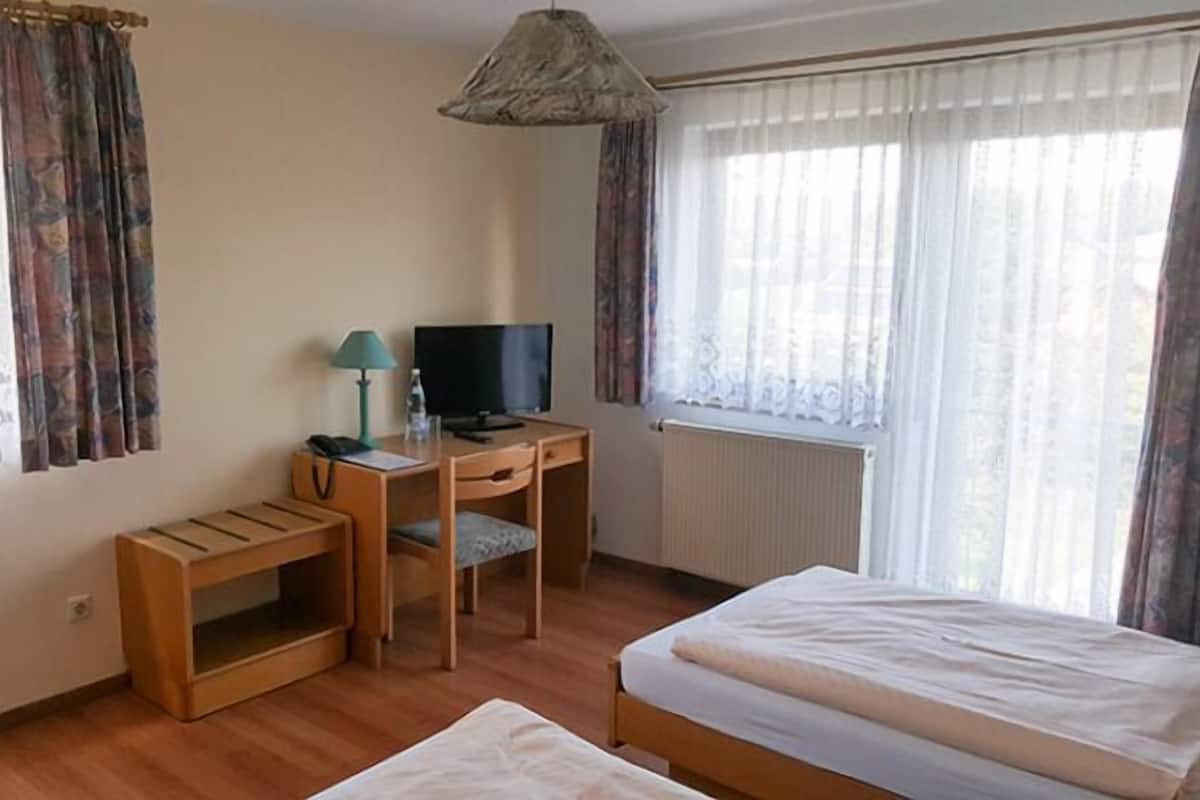 Ein kleiner, ordentlicher Raum mit einem Bett, einem Schreibtisch mit Computer, einer Lampe und einem Fenster mit Vorhängen.
