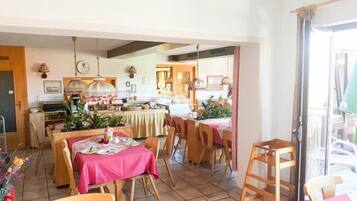 A dining area with tables covered in pink tablecloths, wooden chairs, and a kitchen area with a counter and various items.