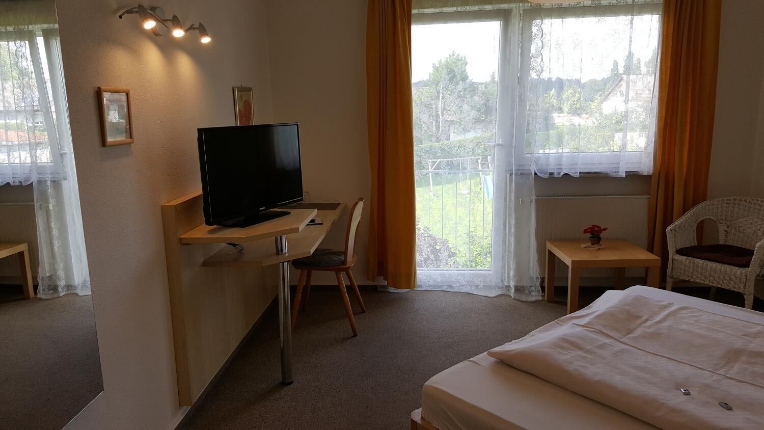 A hotel room with a bed, a desk with a television, a chair, and a small table with a flower vase.