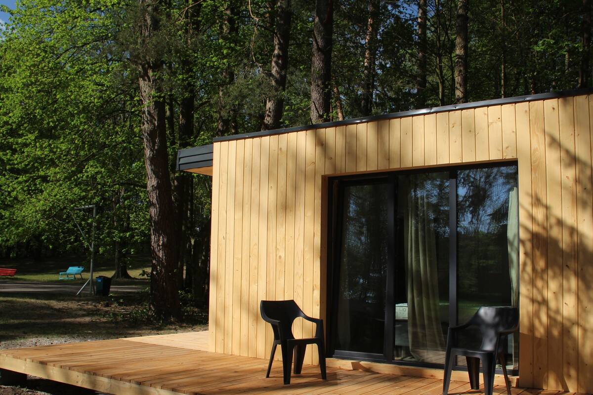 Una cabaña moderna de madera con un amplio ventanal corredizo, una terraza de madera y dos sillas de plástico negro.
