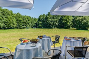 Un área de comedor al aire libre con mesas redondas cubiertas con manteles blancos, rodeada de sillas de mimbre, con vistas a un campo cubierto de hierba y árboles frondosos.