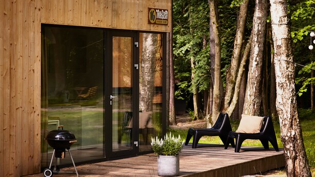 A wooden cabin with a glass door, a barbecue grill, and two black chairs on a deck. - Kaletka (Ilawa)